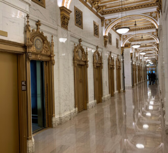 Ornate hallway with gold-trimmed elevators, marble walls, arched ceiling, and polished floors.