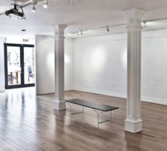 Bright, empty gallery space with wooden floors, white walls, columns, and a black bench in the center.