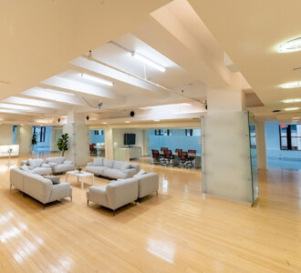 Modern open office with lightwood floors, white sofas, glass-walled meeting room, and bright overhead lighting.