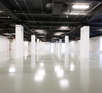 Spacious, empty industrial interior with white columns, polished floor, and large windows on the right.