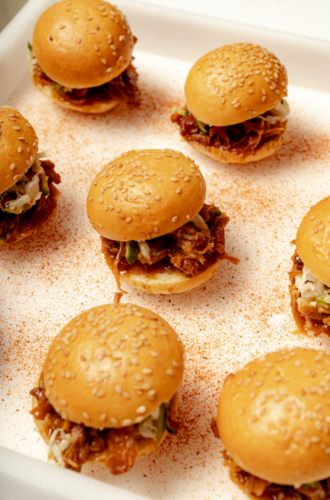 Mini pulled pork sandwiches with sesame buns, perfect for Graduation Catering NYC, arranged on a white tray with seasoning.
