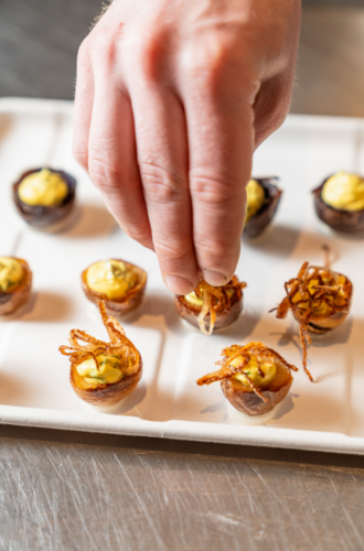 A hand garnishes bite-sized catering appetizers with crispy toppings on a white tray at a Climate Week NYC event.