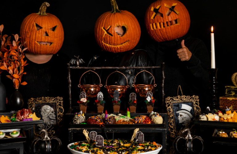 Three people with pumpkin heads stand behind a halloween catering feast, surrounded by spooky foods and eerie decorations.