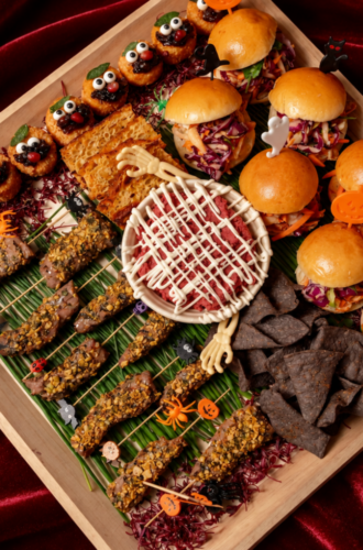 A Halloween catering platter with mini burgers, skewered snacks, a bowl of dip, and black tortilla chips.