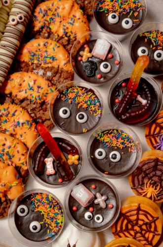 Halloween catering featuring cookies and chocolate pudding cups decorated with candy eyes, sprinkles, and gummy worms.