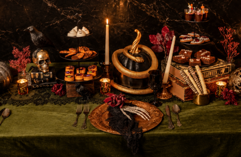 A festive Halloween catering table decorated with skull motifs, a black pumpkin, candles, and tiered trays of treats.